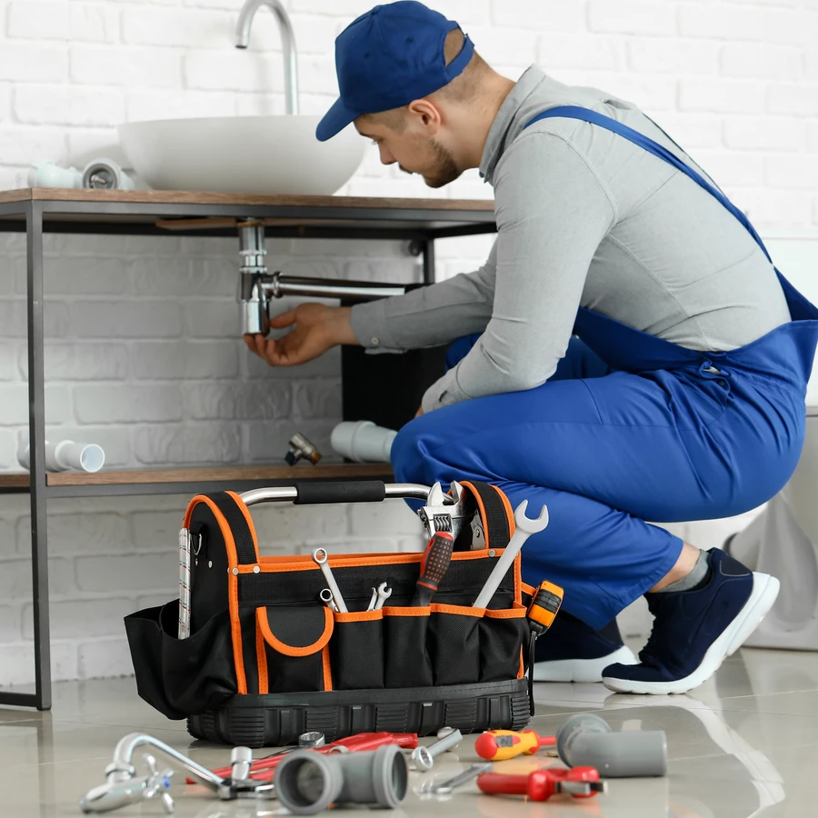 Plombier en uniforme bleu réparant une canalisation sous un lavabo avec sa caisse à outils